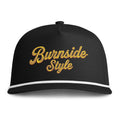 A black baseball cap with 'Burnside Style' embroidered in a textured gold cursive font on the front, featuring a white stripe along the edge of the curved brim.