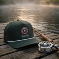 A dark green baseball cap with a 'Real Fly' logo sits on a weathered wooden dock next to a fishing rod and reel, with a misty lake and trees in the background at sunrise.