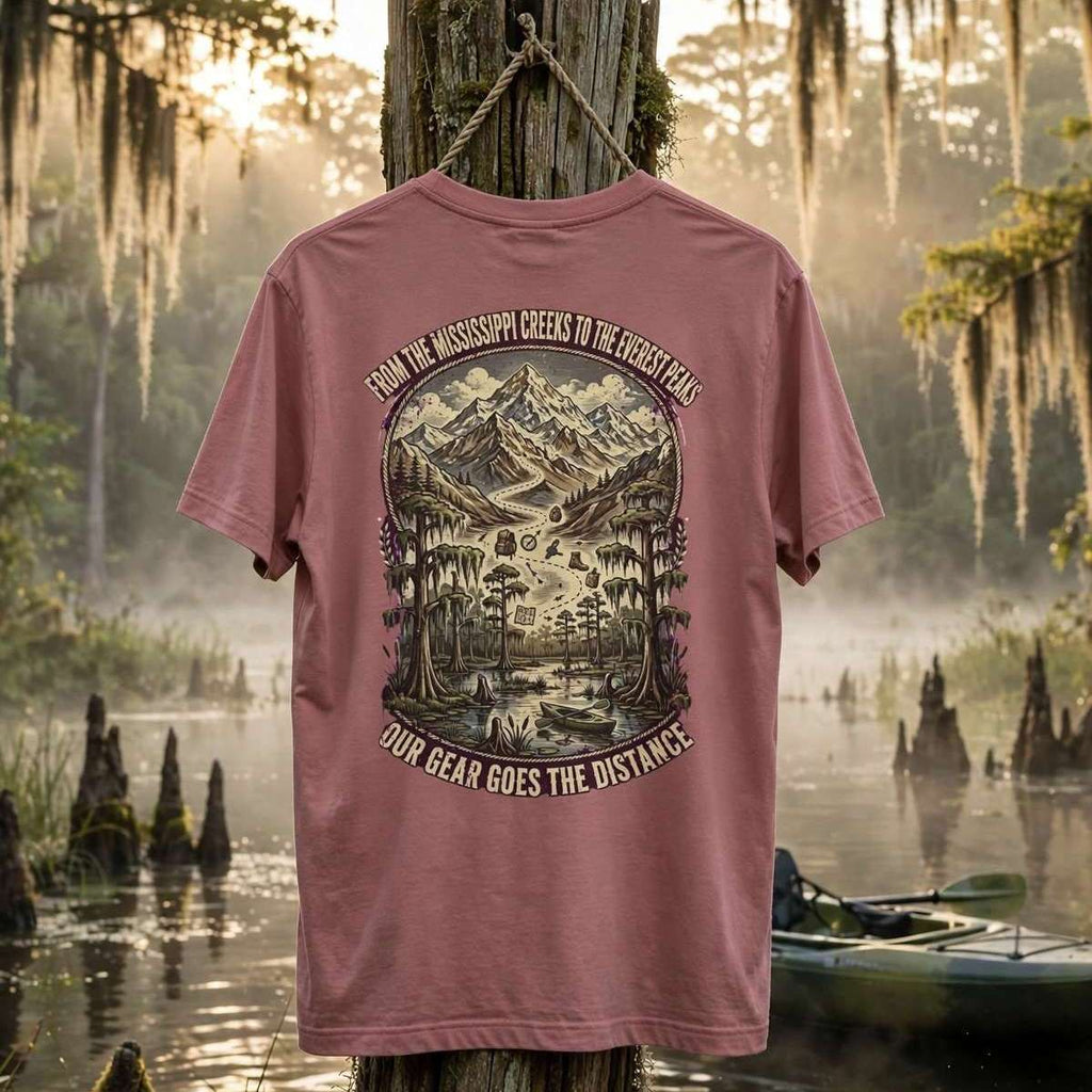 A dusty rose pink graphic t-shirt is displayed hanging from a wooden post in a misty, mossy swamp environment. The back of the shirt features a large detailed graphic of a mountain and forest scene with the text 'FROM THE MISSISSIPPI GREEKS TO THE GREAT BAYES' and 'OUR BEAR GOES THE DISTANCE'.