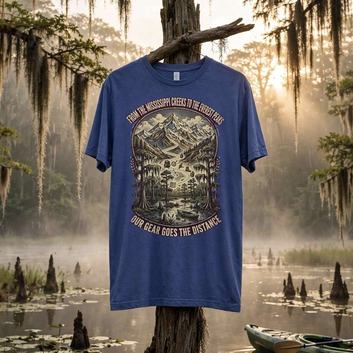 A blue graphic t-shirt hanging from a tree stump in a misty swamp setting with Spanish moss, lily pads, and a green kayak in the water. The shirt features a large front graphic of a mountain landscape with the text 'FROM THE MISSISSIPPI GULF SEES THE GREAT BLUE' and 'OUR BEAR GOES THE DISTANCE'.