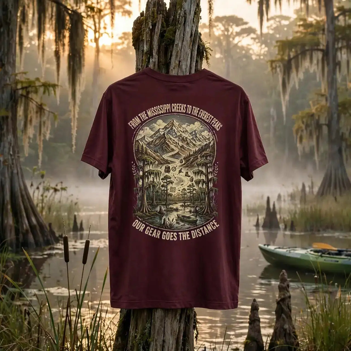A maroon graphic t-shirt is displayed on a weathered wooden post in a misty swamp setting at sunrise. The back of the shirt features a large vintage-style illustration of a mountain and forest, with the text 'FROM THE MISSISSIPPI GUES TO THE COLUMBINE DAY' and 'OUR BEAM GOES THE DISTANCE'.