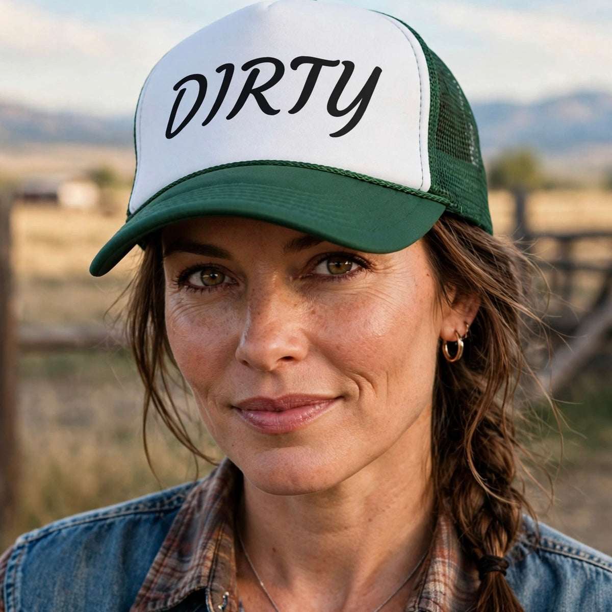 A woman wearing a green and white trucker hat with the word 'DIRTY' printed on the front, photographed outdoors in a rustic rural setting with a fence and mountains in the background.