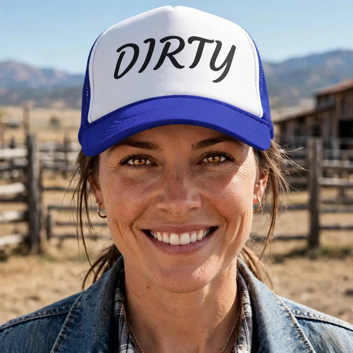 A smiling woman outdoors wearing a white and blue trucker hat with the word 'DIRTY' printed in black across the front, along with a denim jacket and plaid shirt.