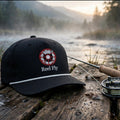 A black baseball cap with a red and white circular logo reading 'Reel Fly' sits on a weathered wooden dock beside a fly fishing rod and reel, with a misty river and forest in the background.
