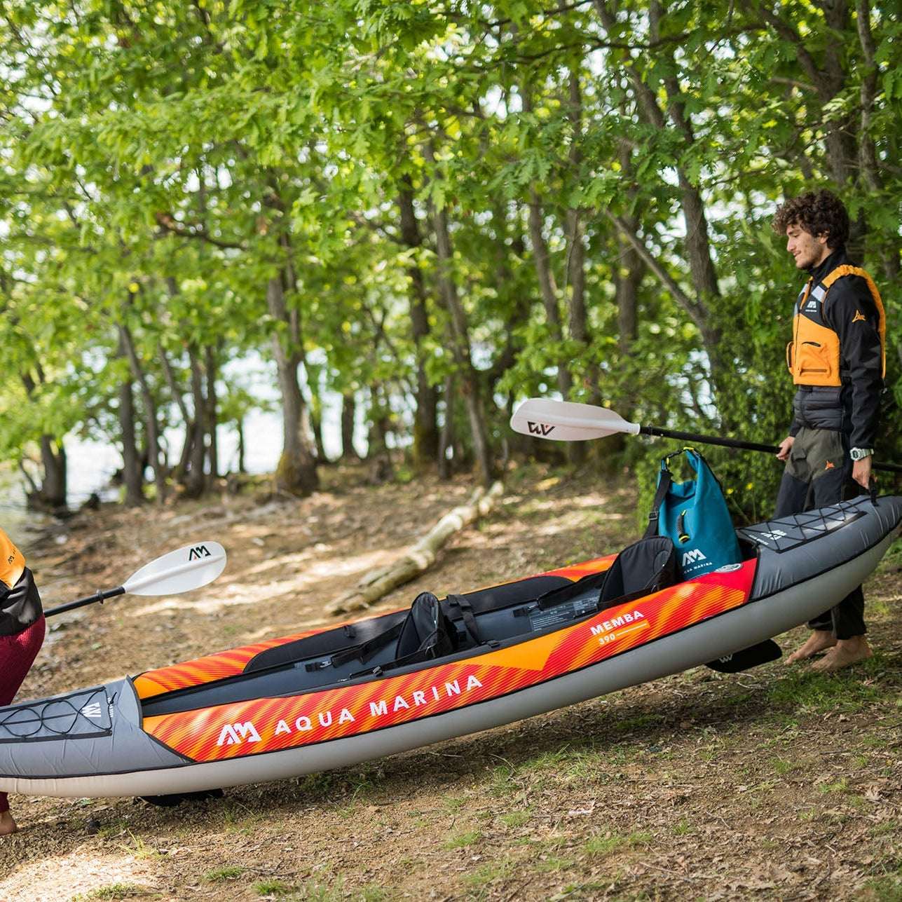 Two people with inflatable kayaks by a lakeside with trees in the background