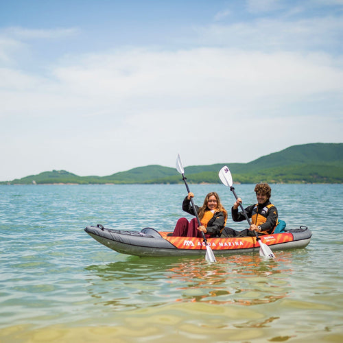 Two people in an inflatable kayak on a calm lake with mountains in the background