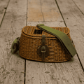 A woven wicker shoulder bag with an olive green fabric strap and a brown leather buckle closure, resting on a rustic wooden plank floor.