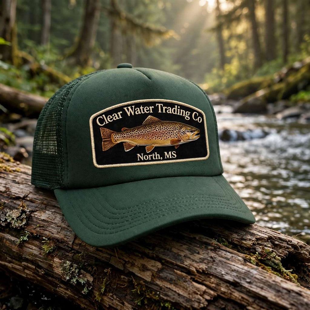 An olive green trucker hat with a 'Clear Water Trading Co' fish patch, resting on a mossy log in a forest stream.
