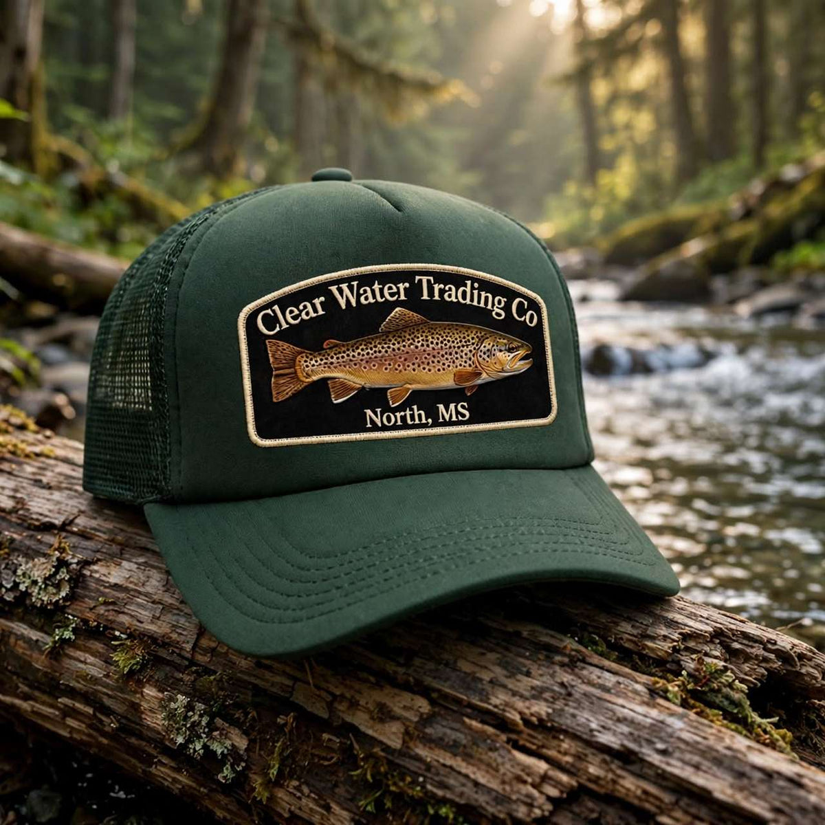 An olive green trucker hat with a 'Clear Water Trading Co' fish patch, resting on a mossy log in a forest stream.