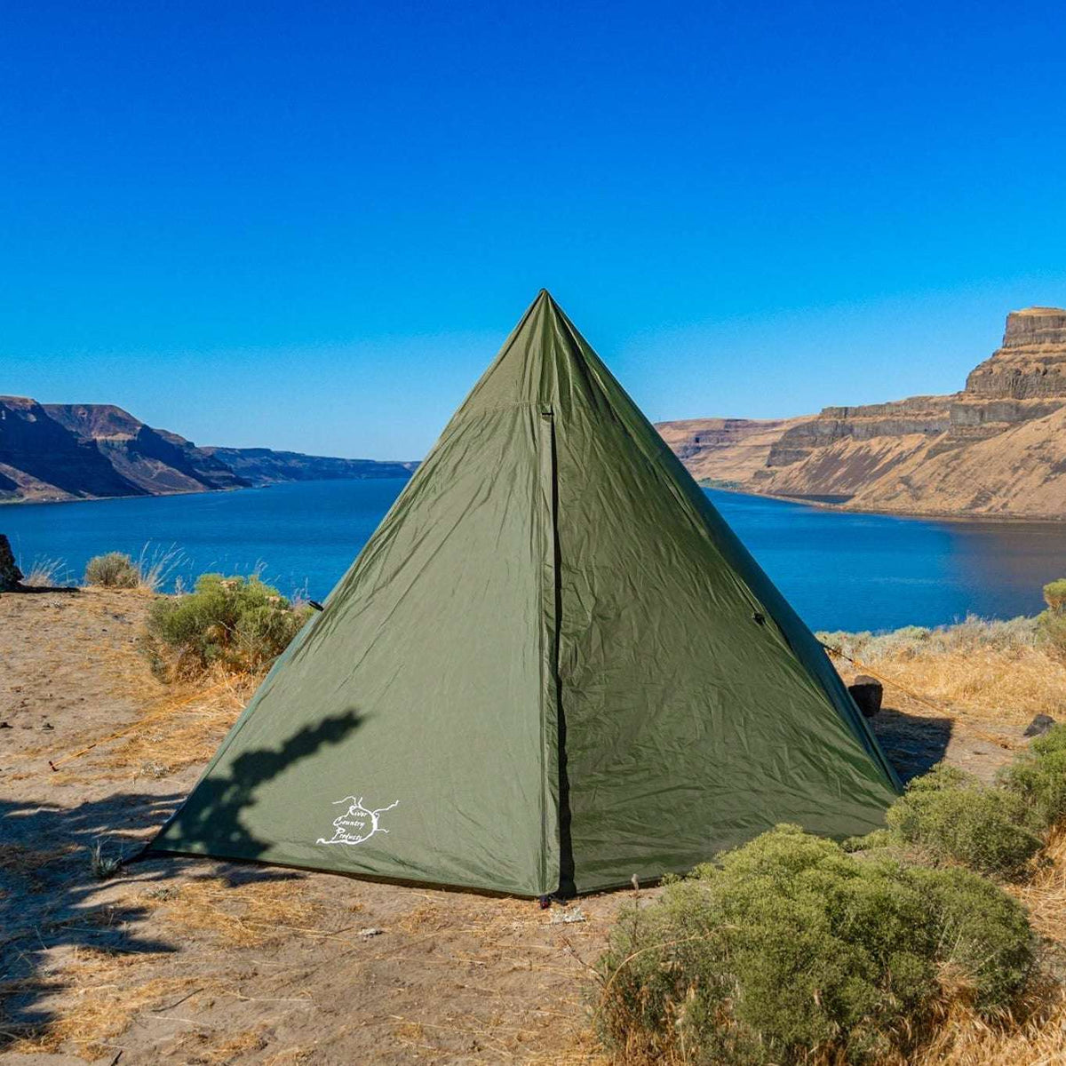 An olive green pyramid-style camping tent pitched on a dry, rocky shoreline with a blue lake and canyon cliffs in the background under a clear blue sky.