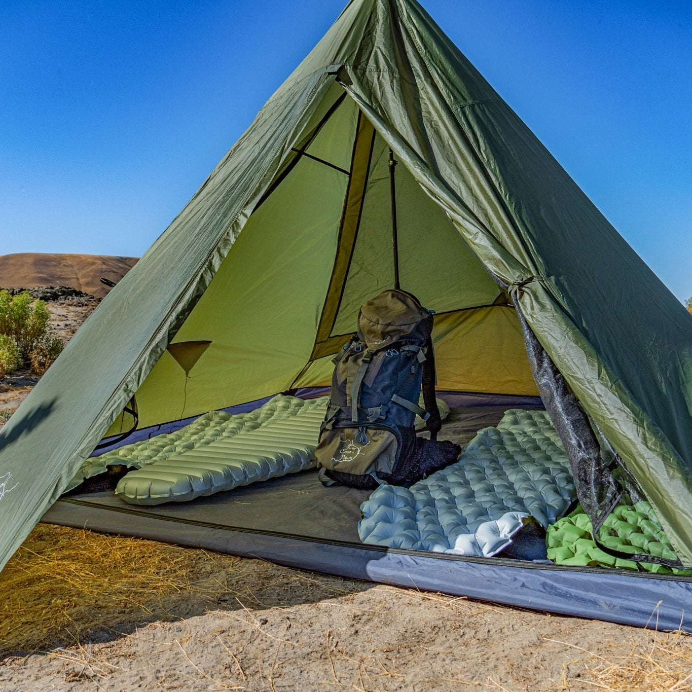 An olive green A-frame camping tent pitched outdoors on sandy ground with a blue sky and water in the background. Inside the tent are sleeping pads, a backpack, and a green sleeping bag.