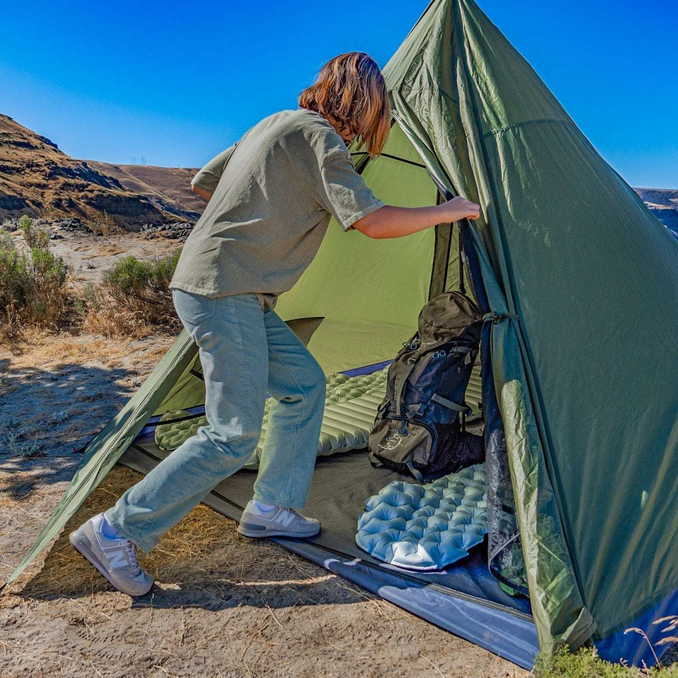 A person sets up an olive green dome camping tent in a dry, mountainous outdoor environment next to a lake under a clear blue sky.
