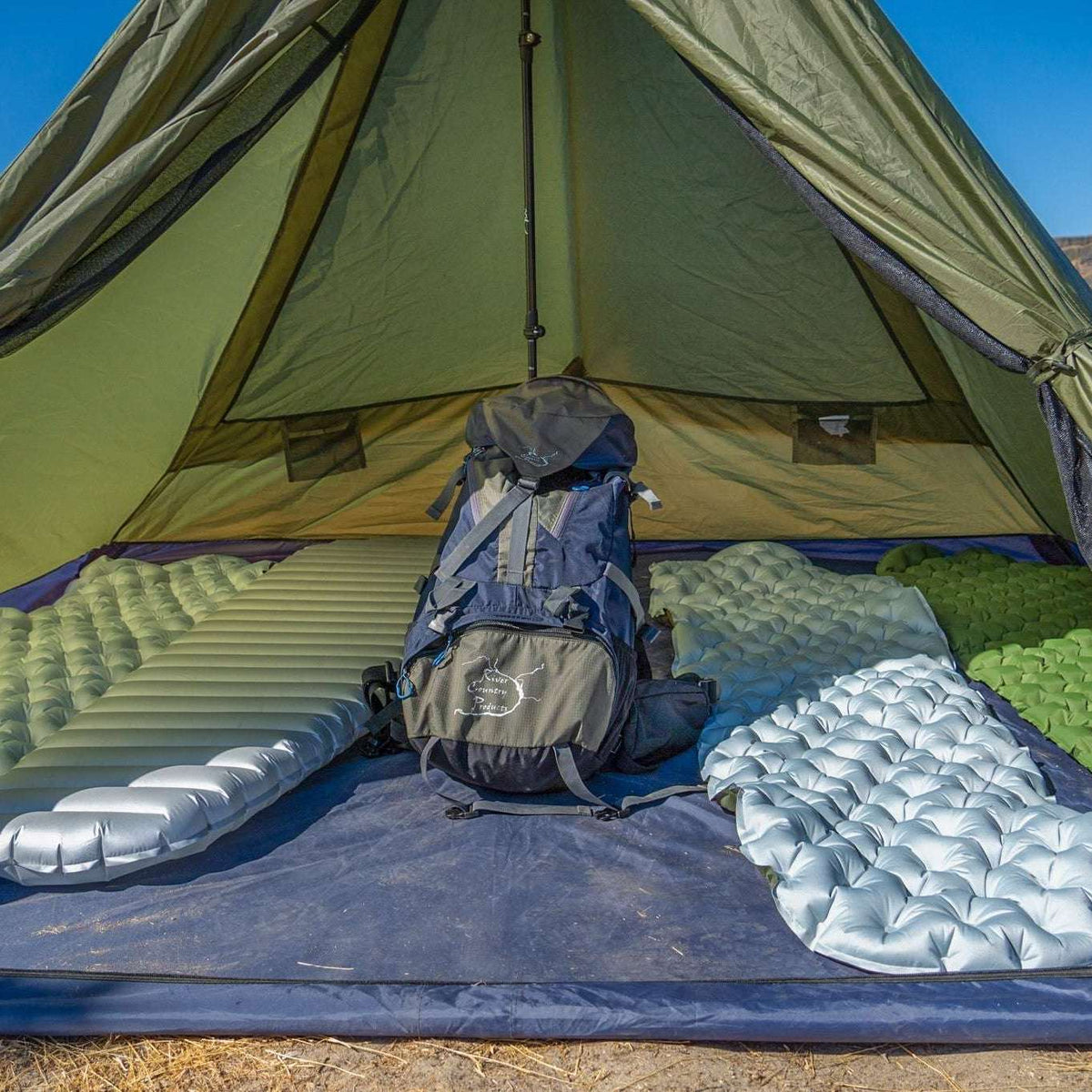 An olive green tent set up in a desert landscape, with camping gear inside including two sleeping pads, a gray backpack, and a blue ground sheet.
