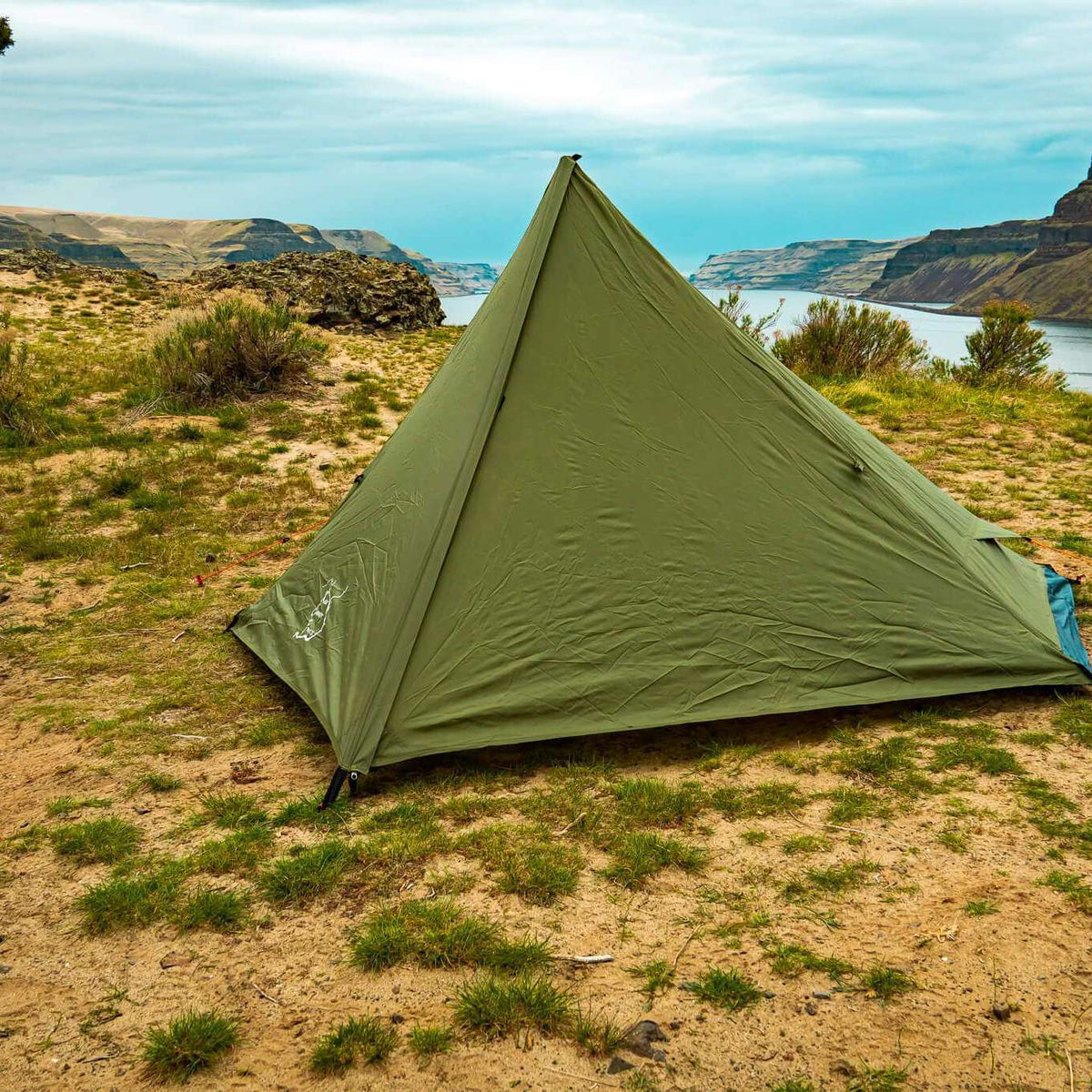 An olive green pyramid-style camping tent pitched on sandy ground with sparse grass, overlooking a canyon and body of water under a cloudy sky.