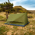 An olive green camping tent pitched on dry grassy ground next to a tree, with rocky hills and a blue sky in the background.