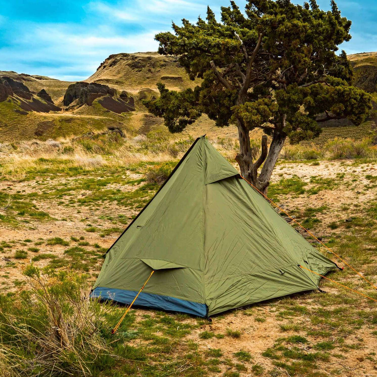 An olive green pyramid-style camping tent pitched on dry grassy ground beside a tree, with rocky hills and a blue sky in the background.