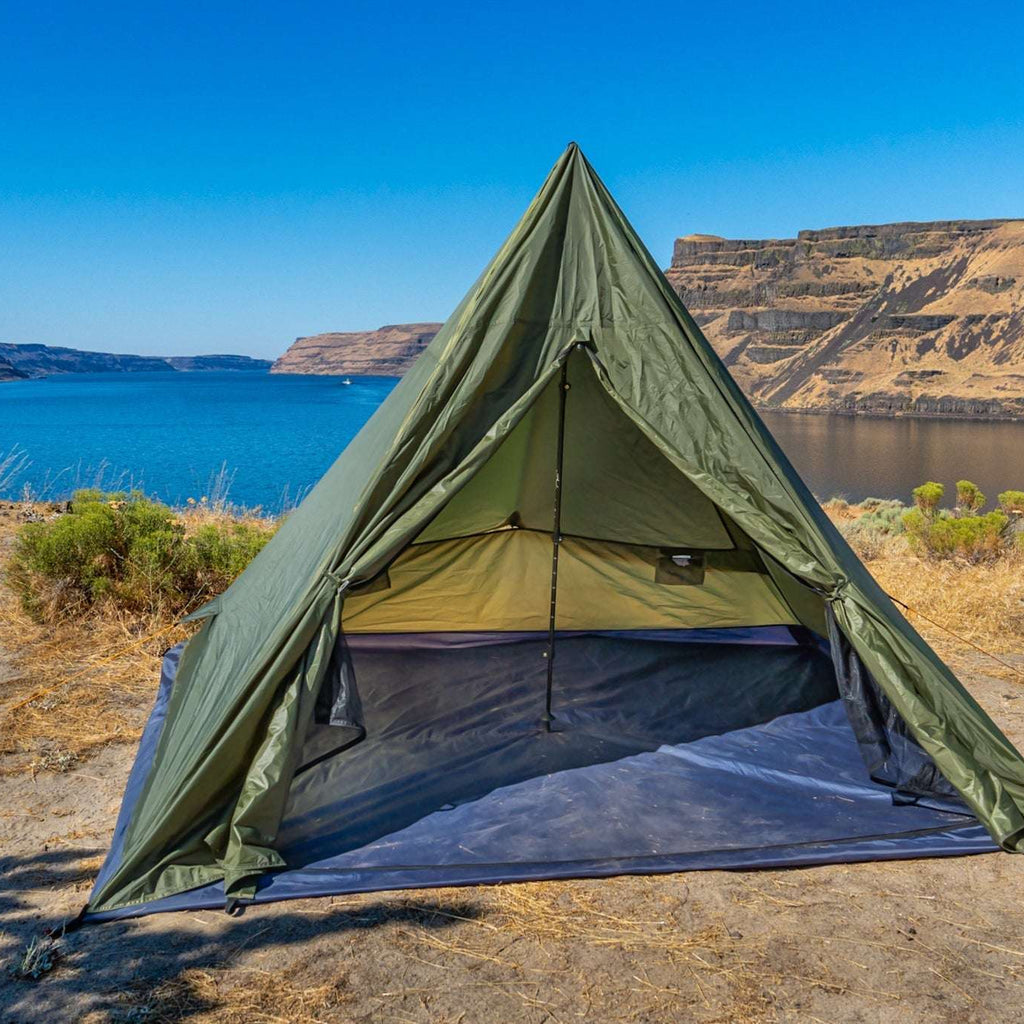 An olive green A-frame camping tent pitched on a dry, sandy lakeshore with a blue lake and canyon cliffs in the background.
