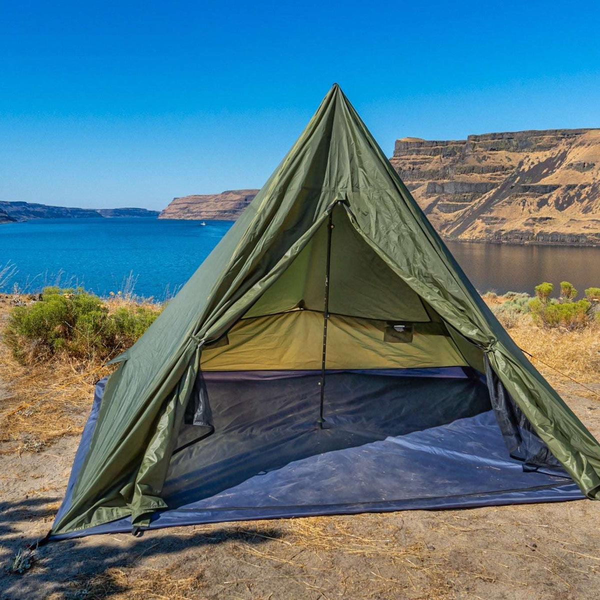 An olive green A-frame camping tent pitched on a dry, sandy lakeshore with a blue lake and canyon cliffs in the background.